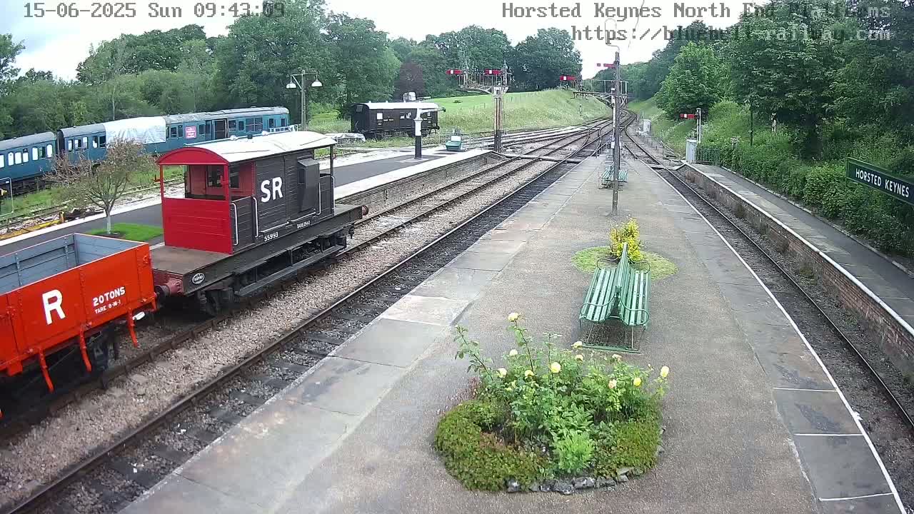 A train station platform with several railway cars, including a red and black engine and a red flatbed car, under an overcast sky.