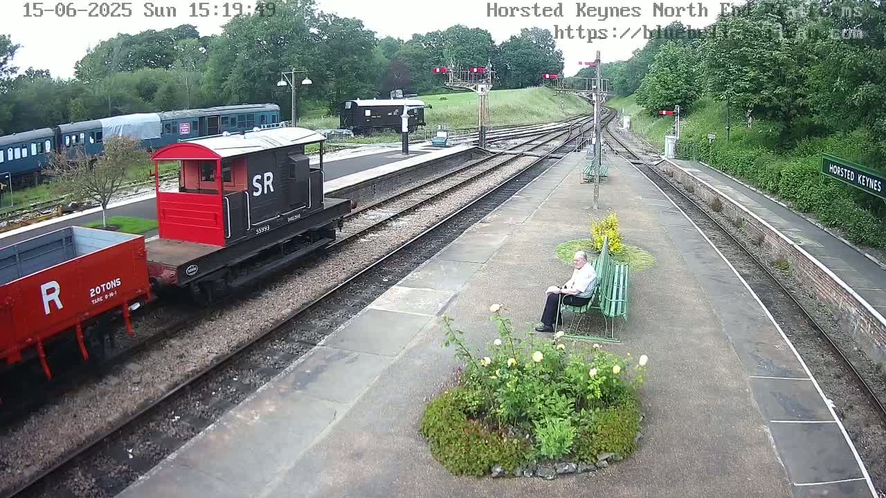 A man sits on a bench at a train station on a partly cloudy day, with several railcars and a passenger train visible nearby.