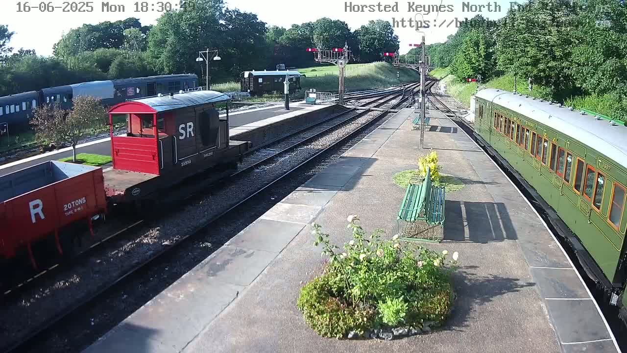A sunny train station with several railway cars, including a red and black railway brake van, a dark-colored passenger car, and an olive-green passenger coach on the tracks.