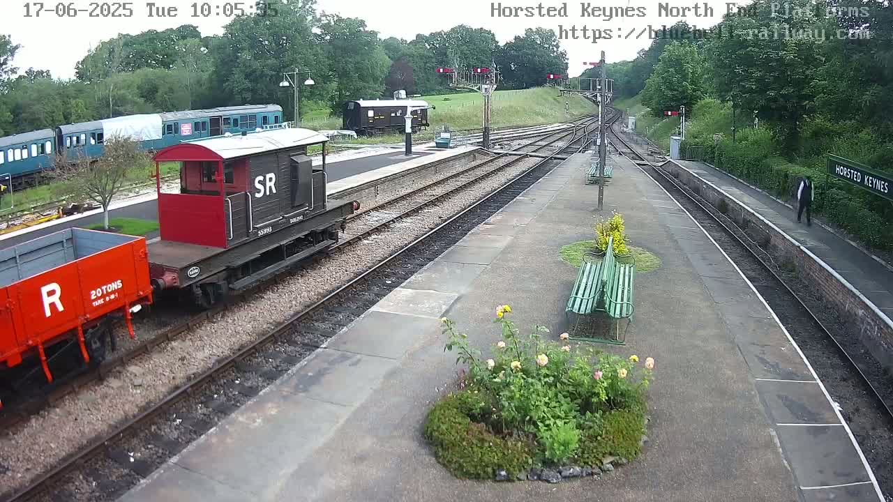 A railway platform with a red and black railway carriage and a red wagon, alongside a train and several benches, under a partly cloudy sky.