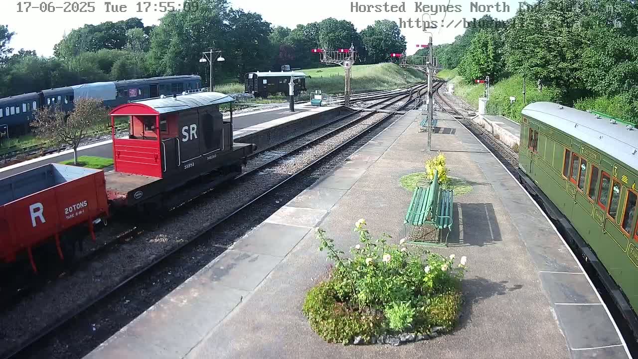 A sunny day at a train station shows several railway cars, including a red and black engine, a green passenger car, and a red railcar, along with a small plant bed near a bench.