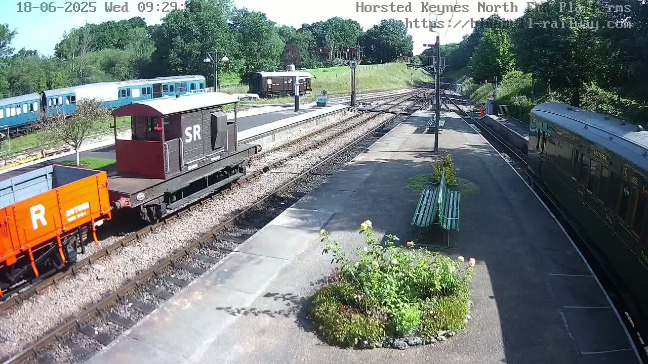 A sunny day at a train station shows multiple railway cars and locomotives on tracks, with a small garden in the foreground.