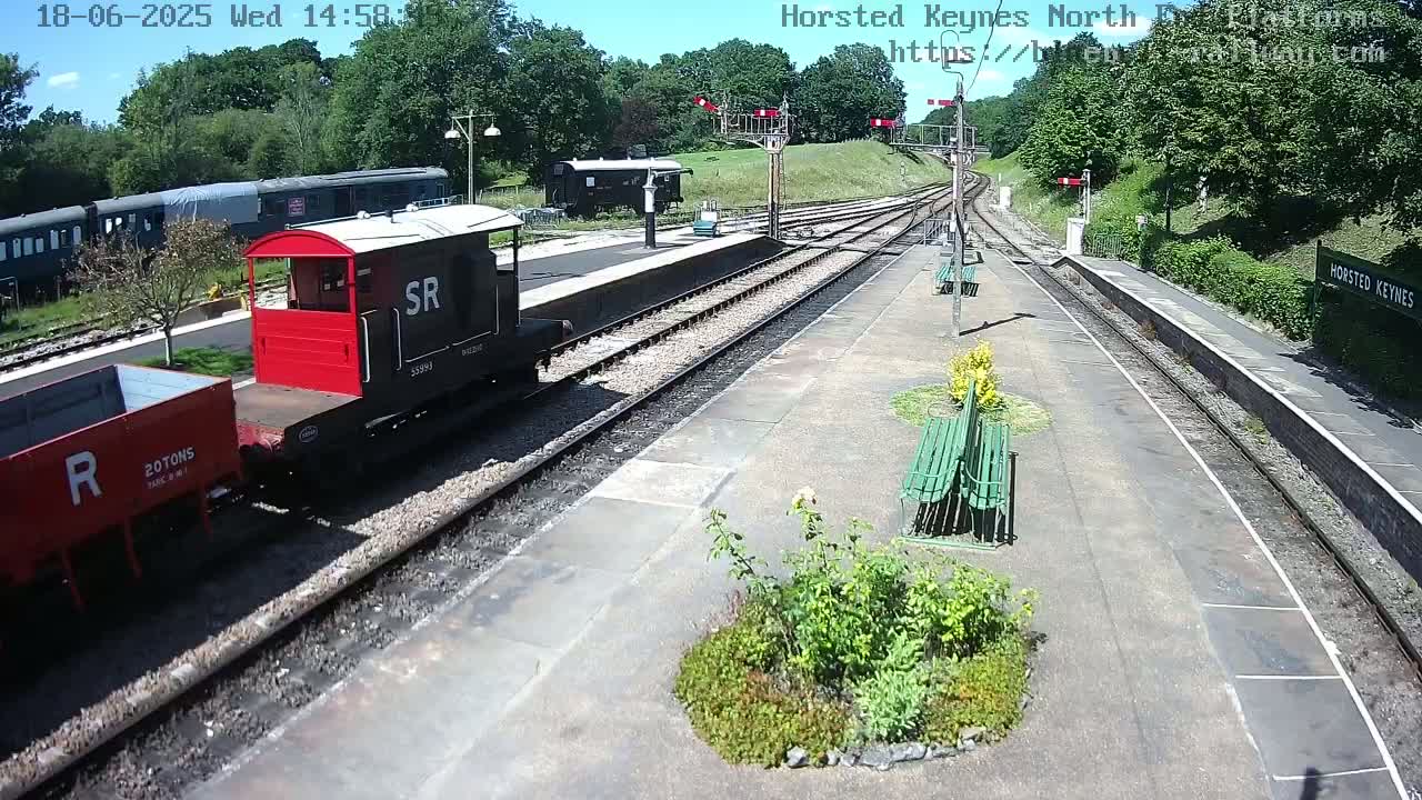 A sunny day at a train station shows a red and black railway carriage and a dark gray freight car on the tracks, with a dark passenger train visible in the background.