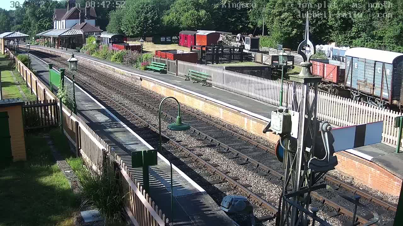 A sunny day at a railway station with multiple train cars and tracks, a station building, and wooden fencing.