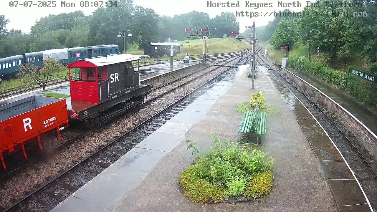 A rain-soaked railway station platform features several railway cars, including a red and black boxcar, and a red flatcar, with several empty train tracks and green benches visible.