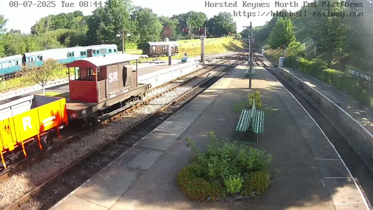 A sunny train station with railway tracks, several train cars, and green benches.