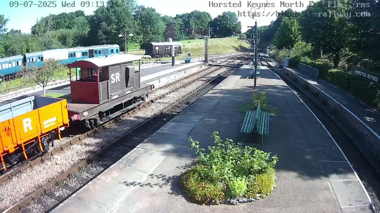A sunny day at a train station shows a railway with several train cars, including an orange and gray boxcar and a red and brown boxcar, parked along the platform.