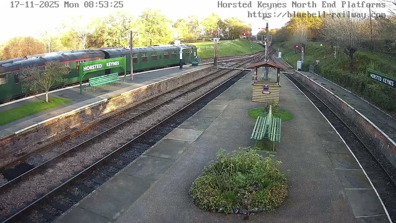 A green passenger train is stopped at a multi-platform train station featuring green benches and a wooden shelter, surrounded by autumn trees under clear, sunny daytime conditions.