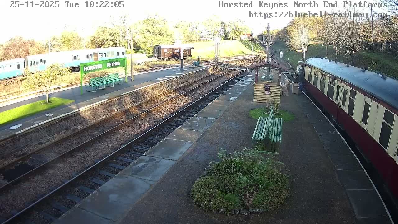 On a bright, sunny day, two trains are visible at Horsted Keynes station, with a light blue train on the far platform and a red and cream train on the near platform, amidst multiple tracks, green benches, and autumn trees.