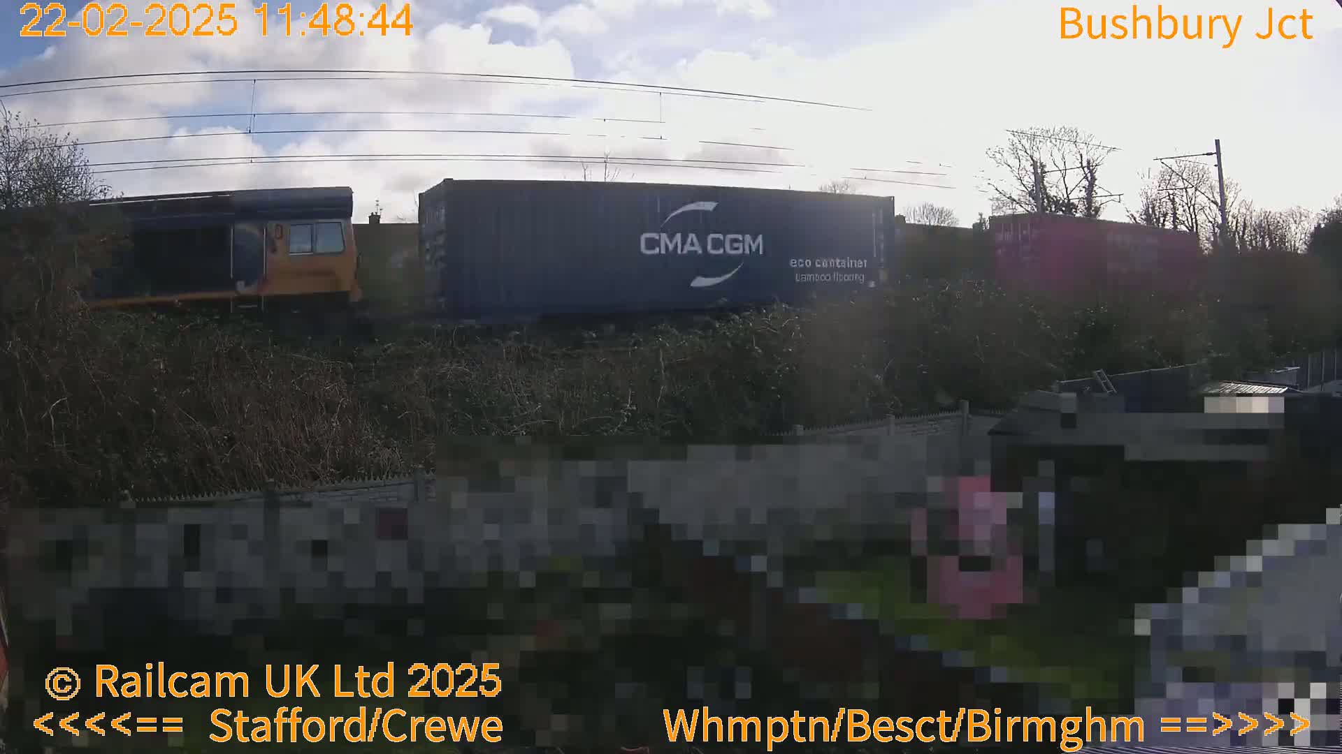 A freight train, partially obscured by bushes, passes under a partly cloudy sky.