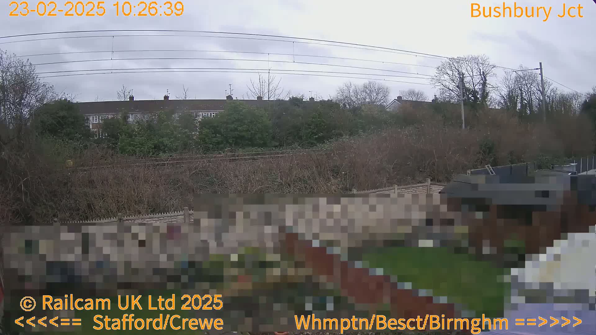 A view of a residential area with overcast skies, showing a garden, a fence, and railway lines beyond it.