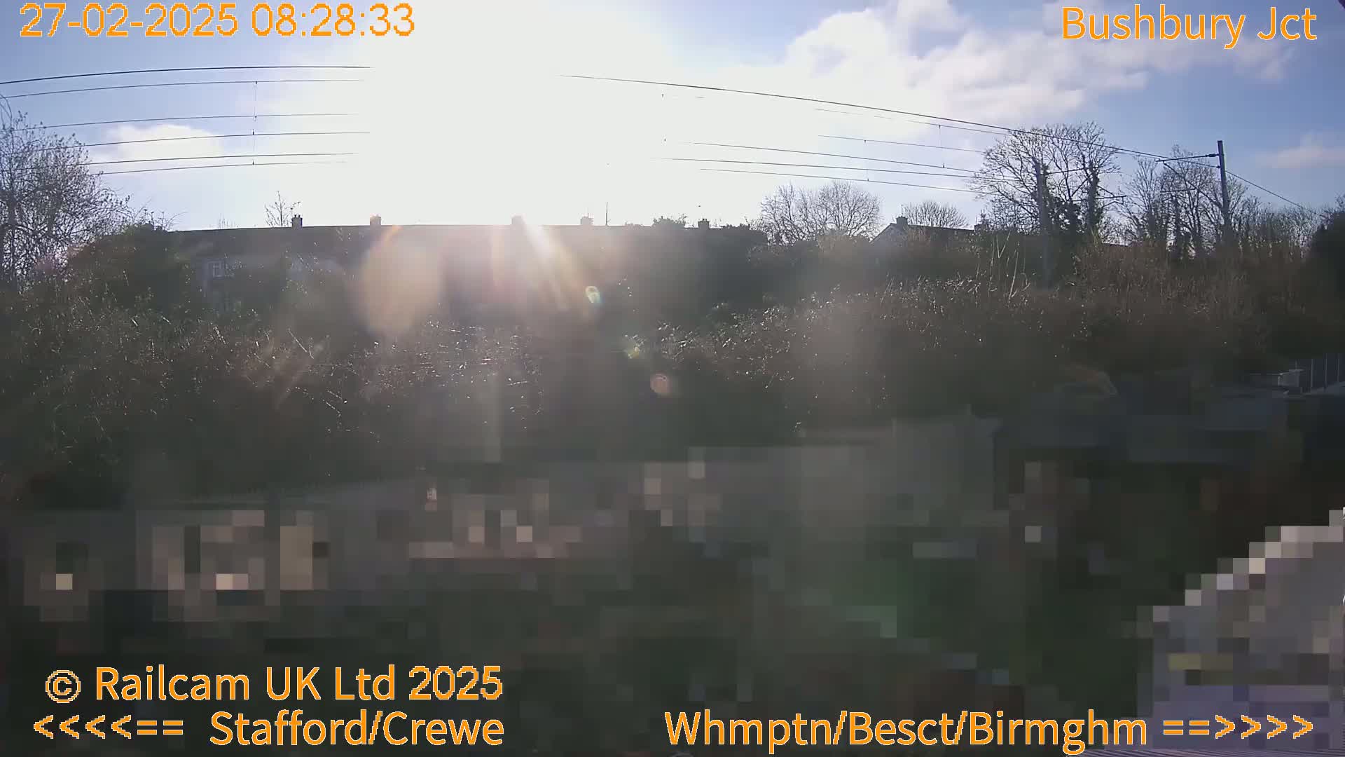 A sunny day reveals a view of rooftops and trees behind a fence, with power lines overhead.