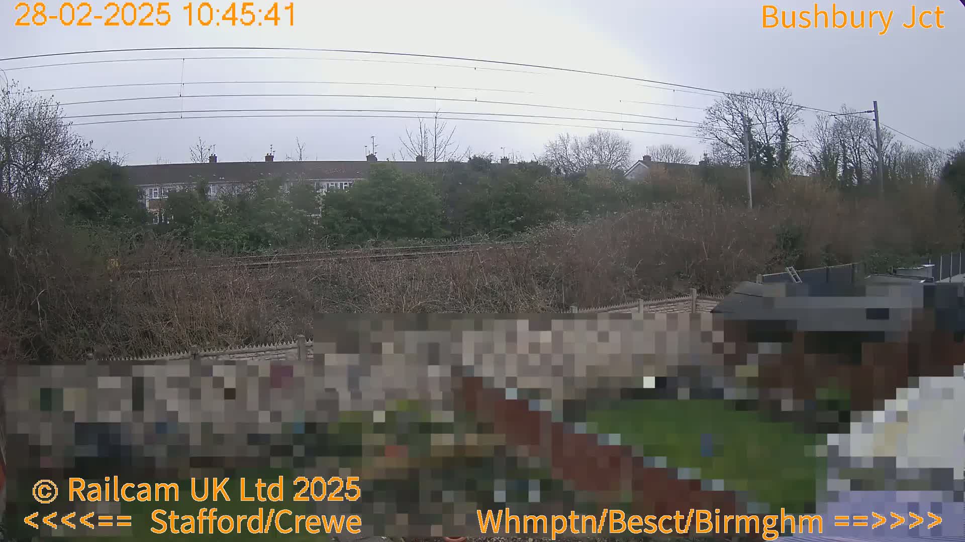 A view of an overcast day shows a residential area beyond a railway line and a garden with a brick wall.