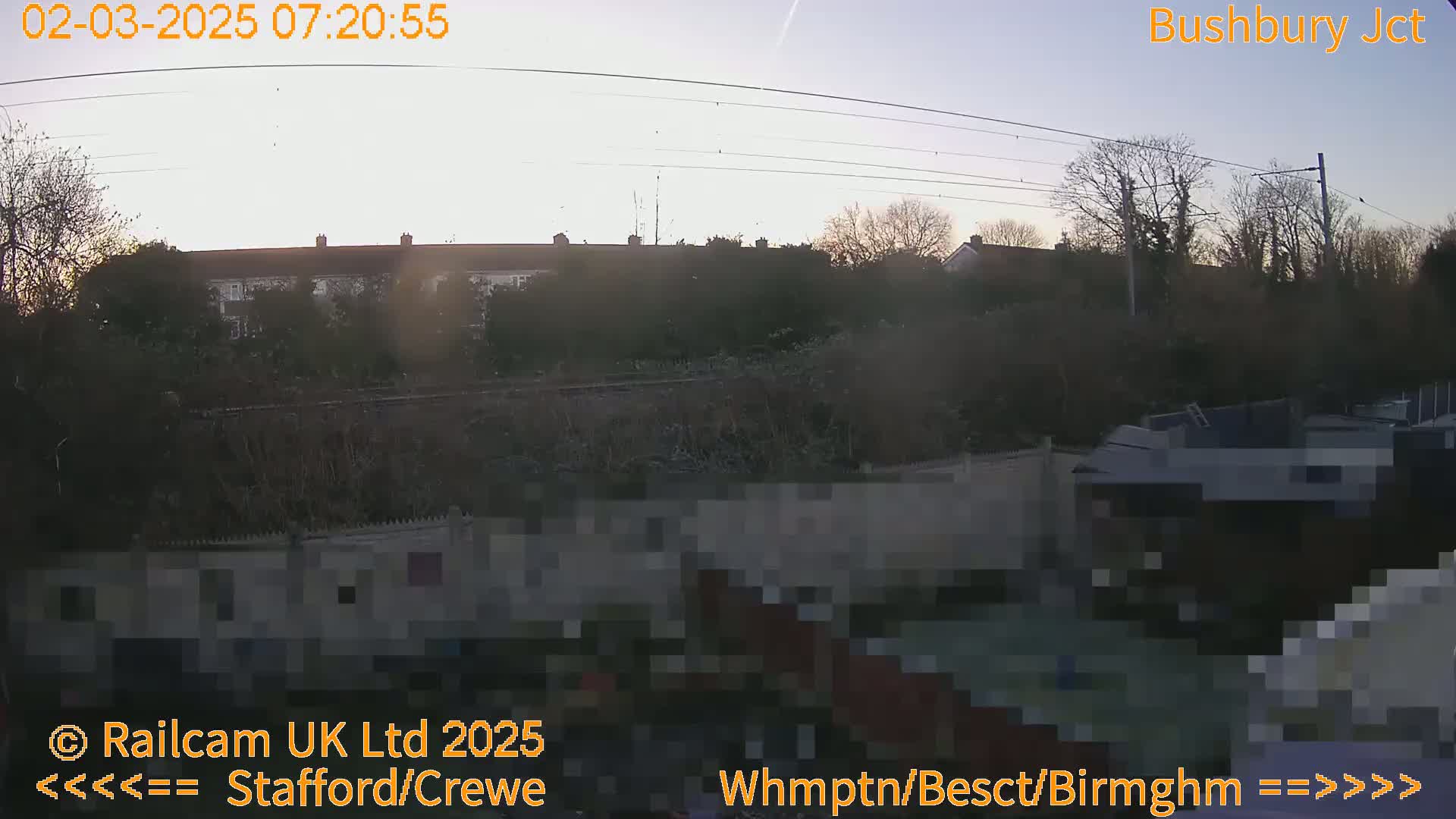 A hazy sunrise illuminates a residential area behind a fence and a railway line.