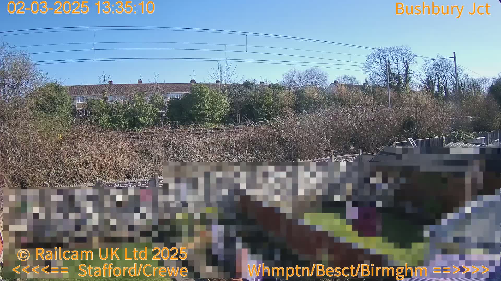 A sunny view of a residential area behind a garden fence and railway lines, with leafless trees and shrubs.