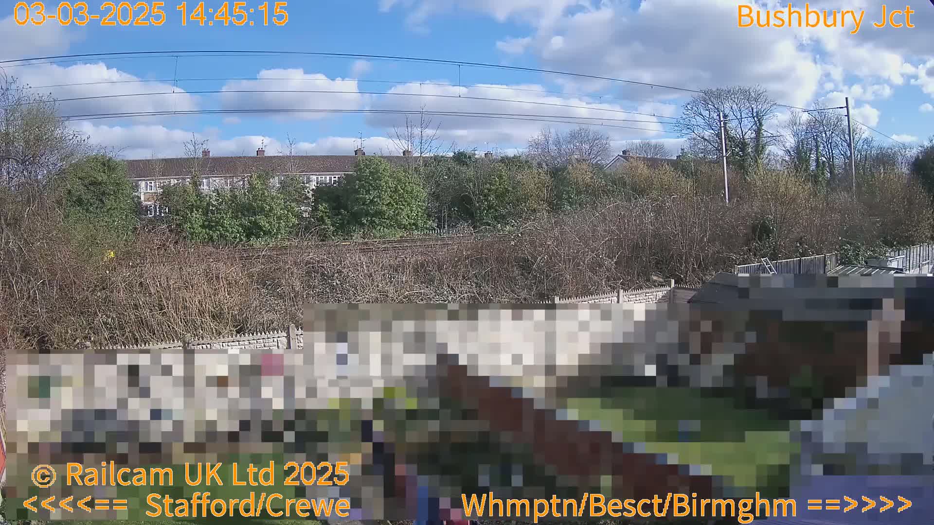 A view of a backyard with a partially visible fence and shed, overlooking a railway line and residential buildings under a partly cloudy sky.