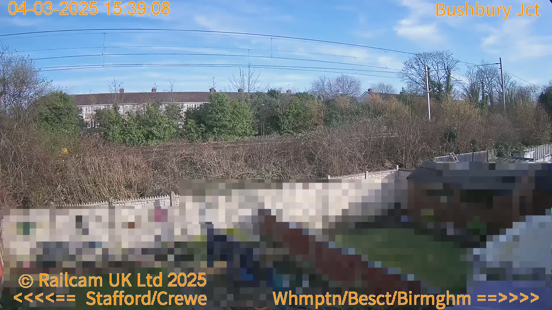 A sunny view of a residential area with overhead power lines, trees, and a backyard containing a shed, seen from across a fence and railway line.