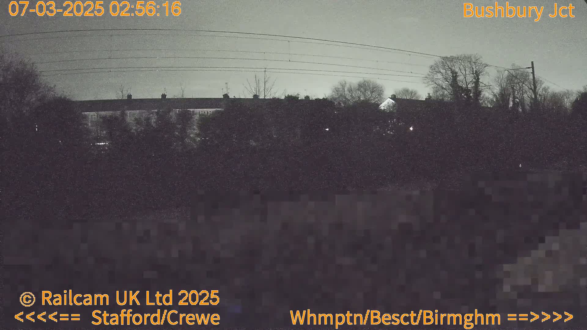 A nighttime, low-light view of residential buildings partially obscured by trees and overhead power lines.