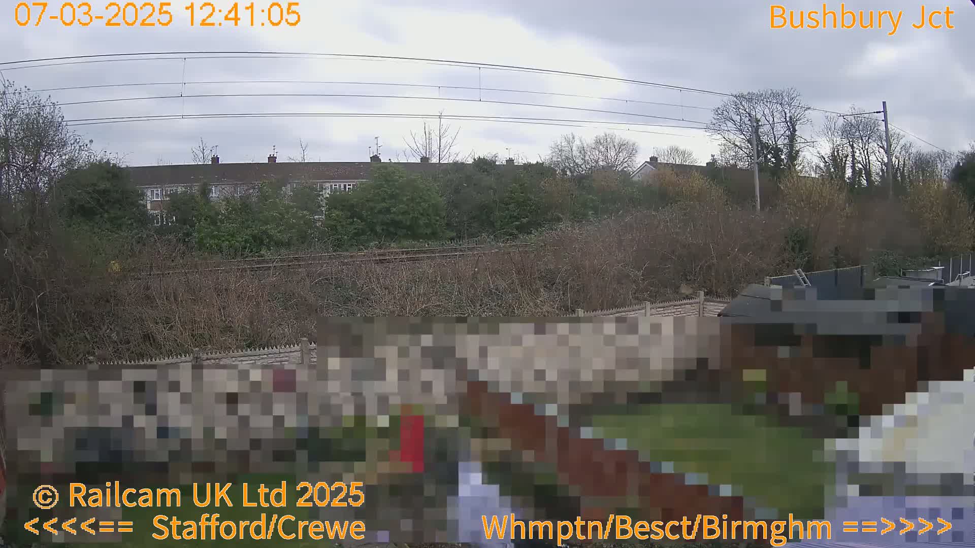 A view of overcast skies over a residential area with a railway line in the distance, partially obscured by shrubs and trees.