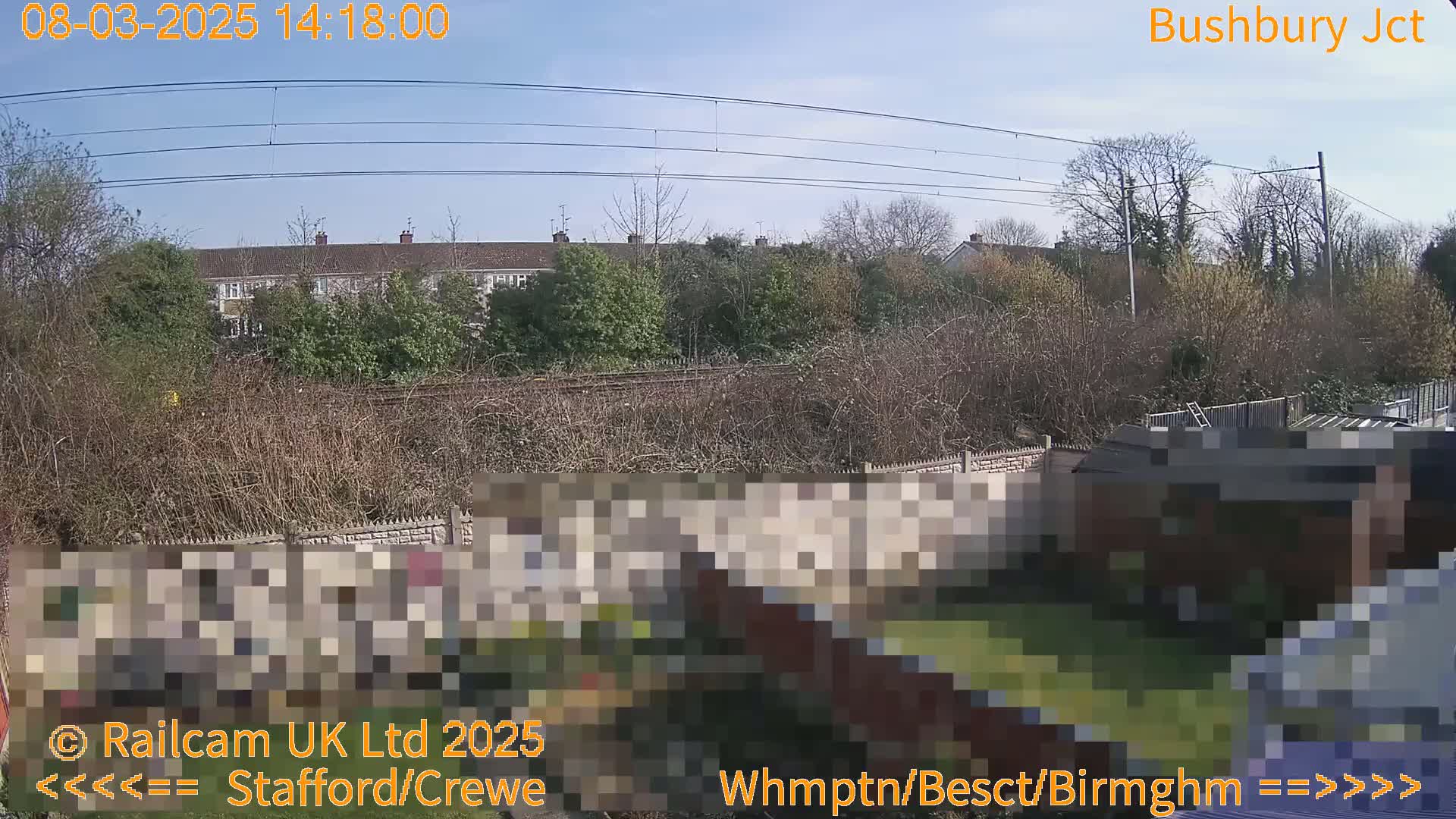 A residential backyard with a partially obscured shed, overlooked by railway lines and houses under a clear, sunny sky.