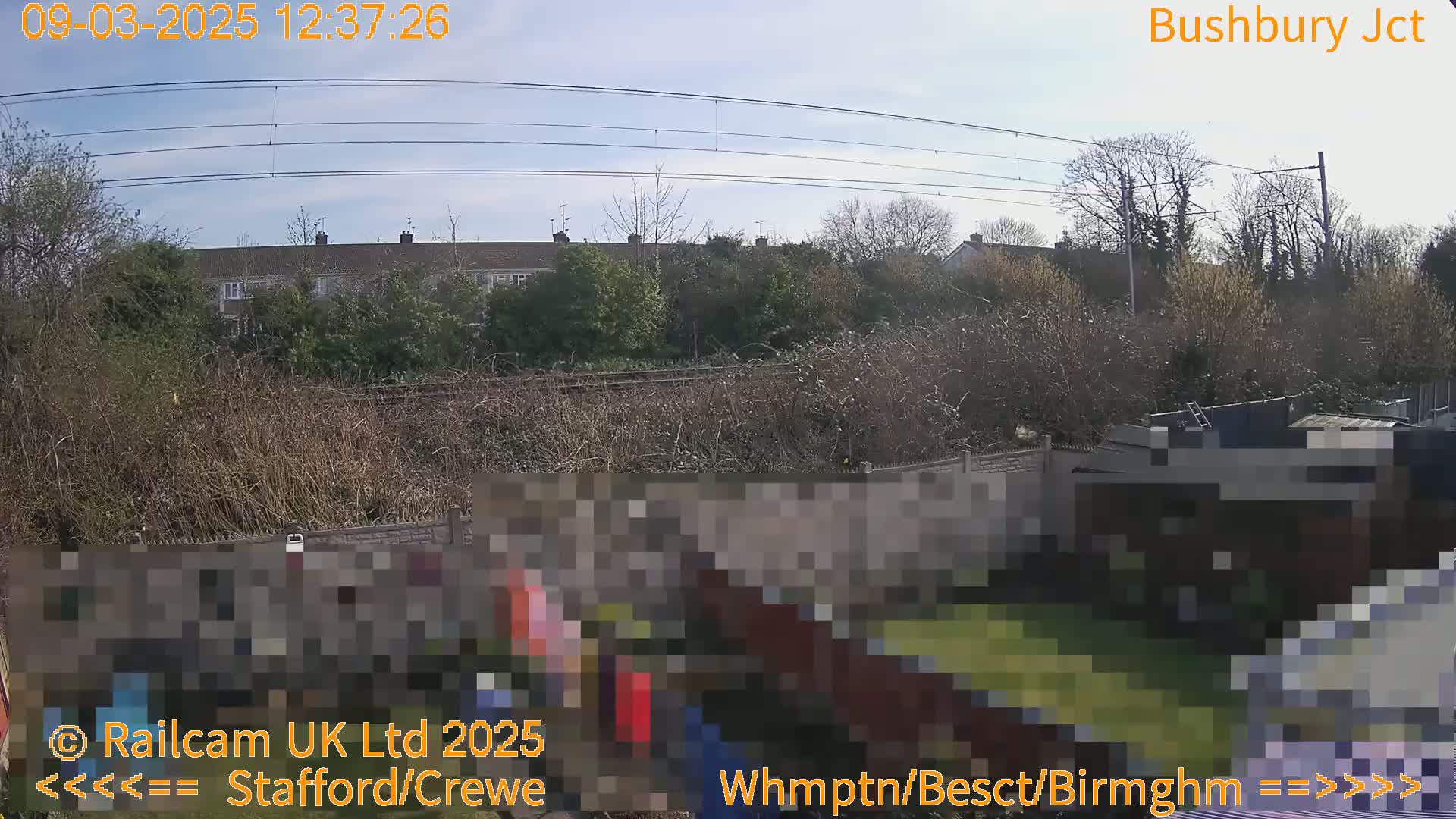 A sunny outdoor view of a railway line behind a garden fence and houses.