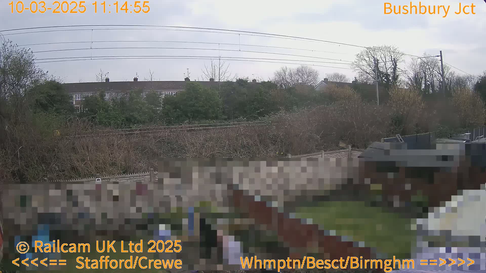 A view of a residential area with a garden, railway tracks, and overhead power lines under an overcast sky.