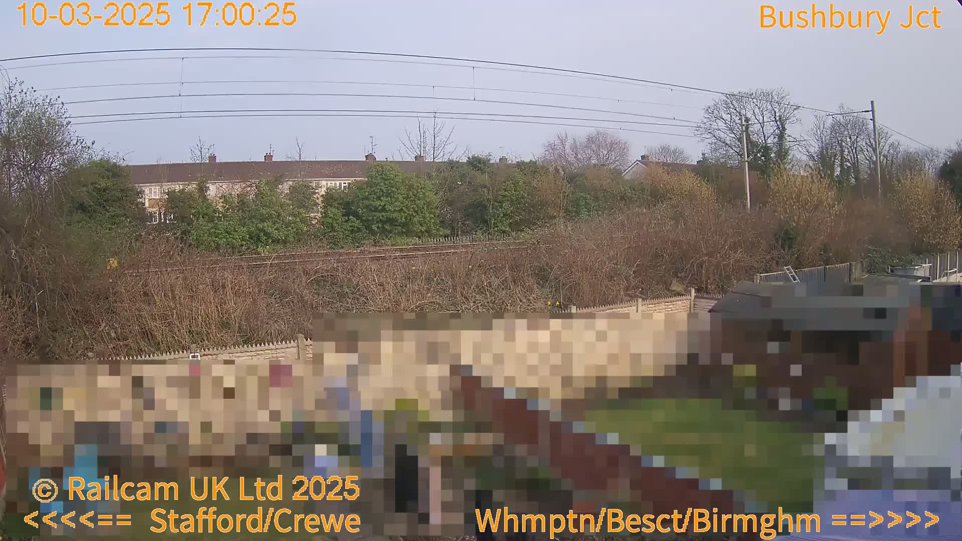 A backyard scene with a fence, a shed, and a grassy area, overlooked by a row of houses and railway lines under a clear sky.