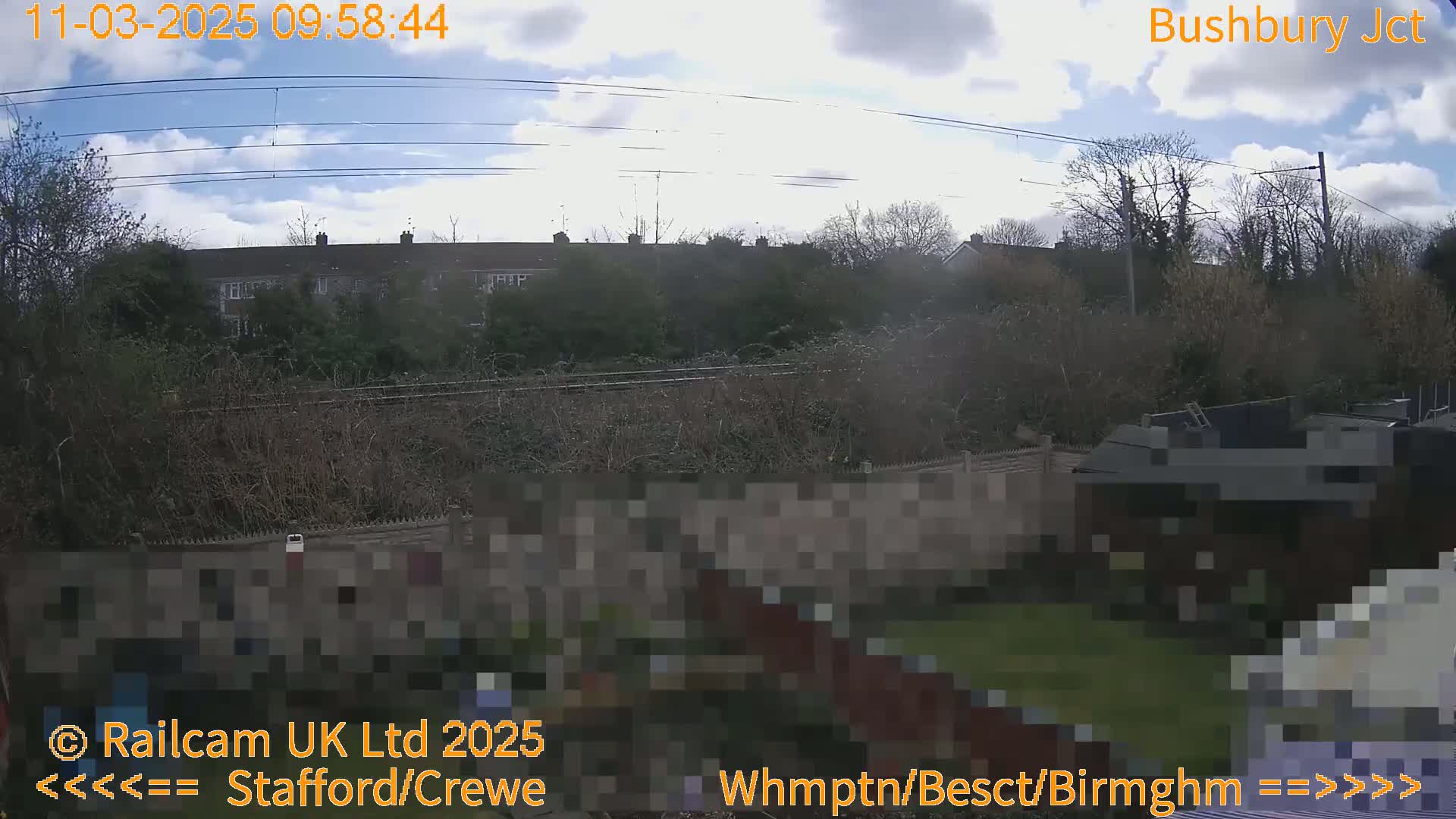 A partly cloudy sky is visible over a residential area with a railway line and vegetation in the foreground, viewed from a distance.