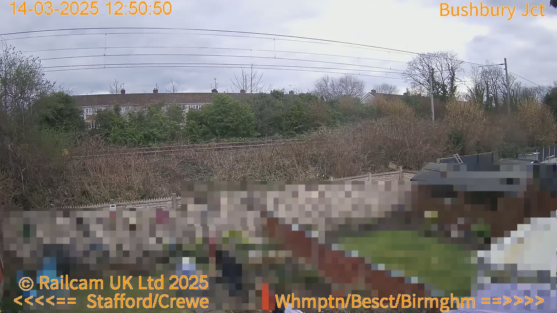 A view of a backyard garden next to a railway line under an overcast sky shows houses and trees in the background.