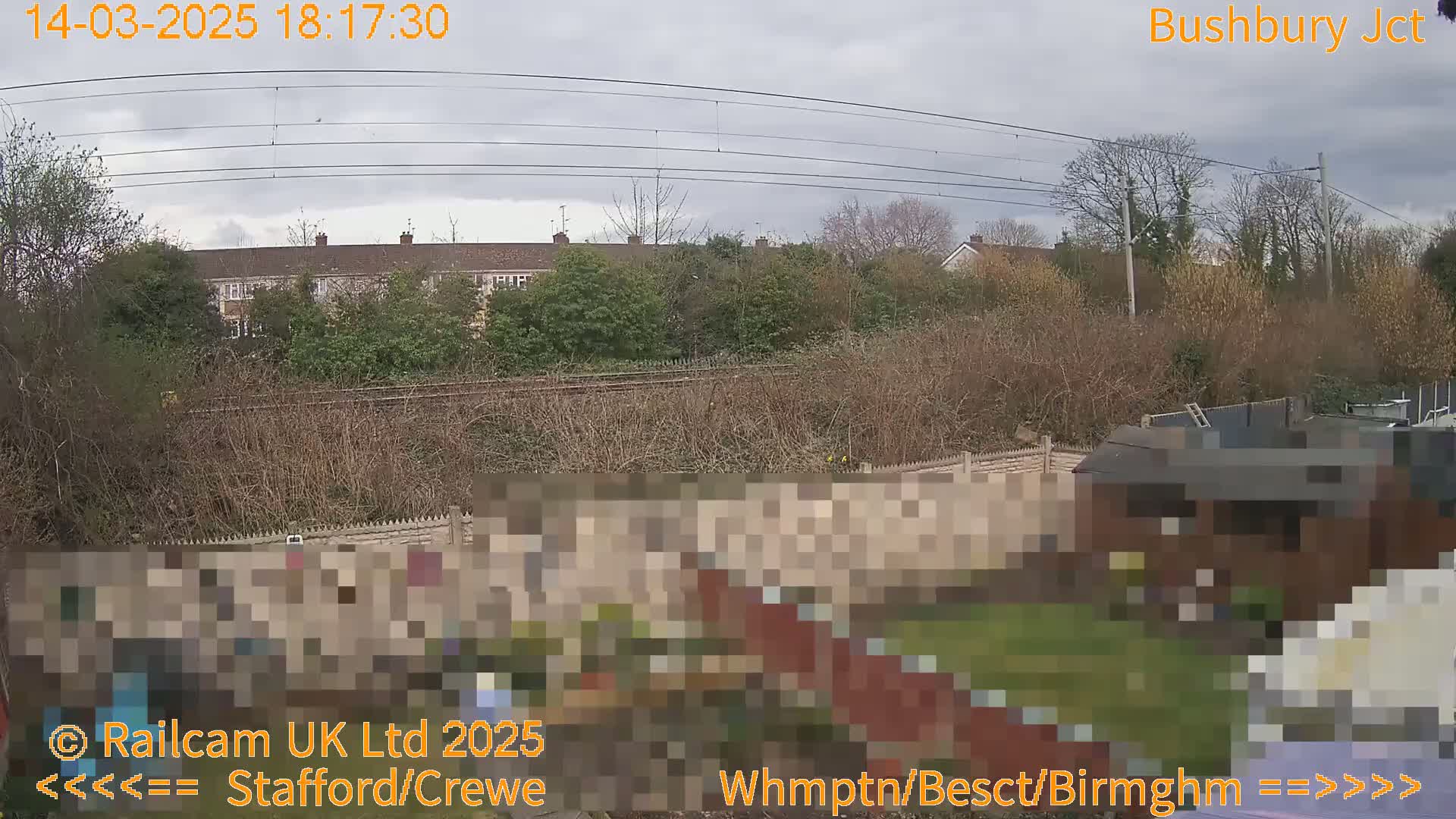 A view of a backyard with a fence, garden, and shed, overlooking railway tracks and a row of houses under a cloudy sky.