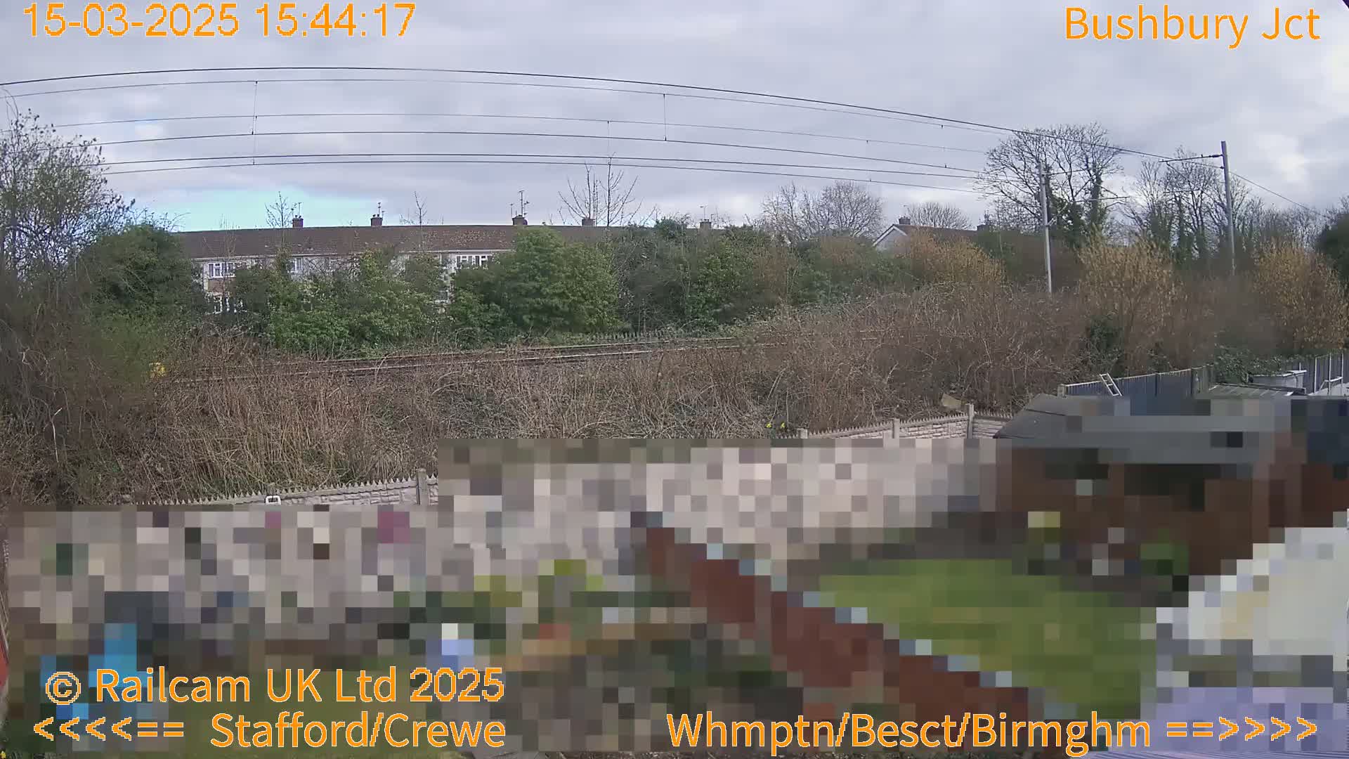 A backyard with a fence and shed is shown, overlooking a railway line and a row of houses under a cloudy sky.