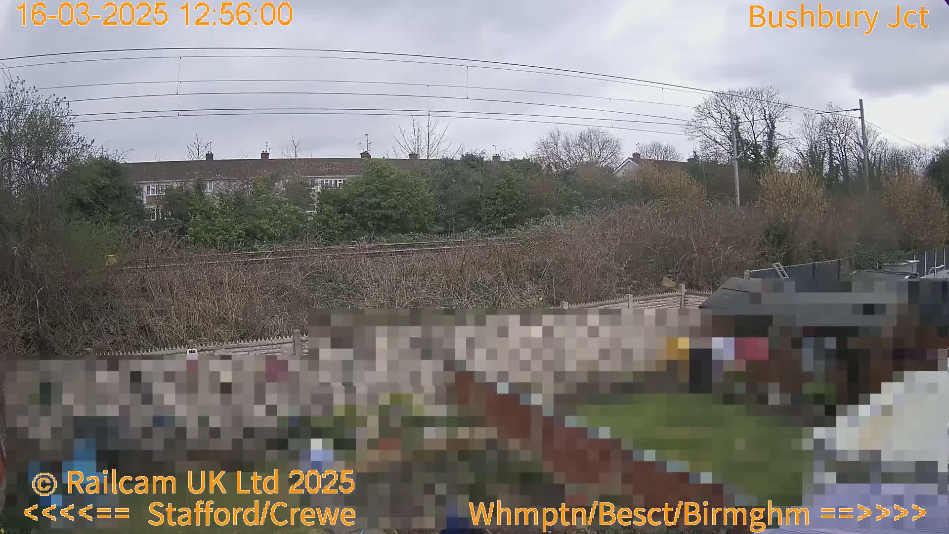 A view of a residential area with overcast skies, showing a backyard garden next to a railway line and several buildings in the background.