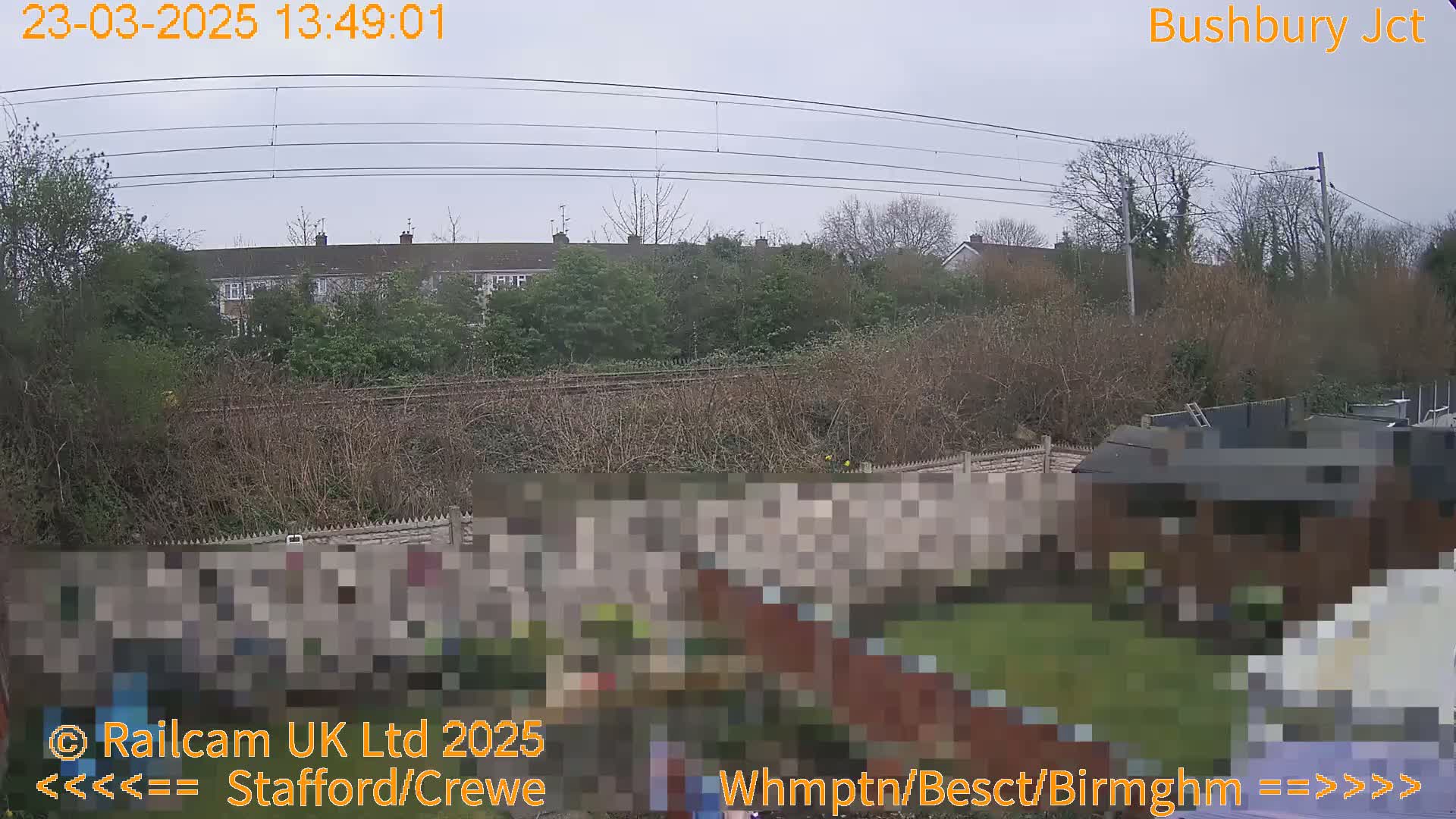 A view of a backyard with a fence, overgrown shrubs, and a building behind a railway line under an overcast sky.
