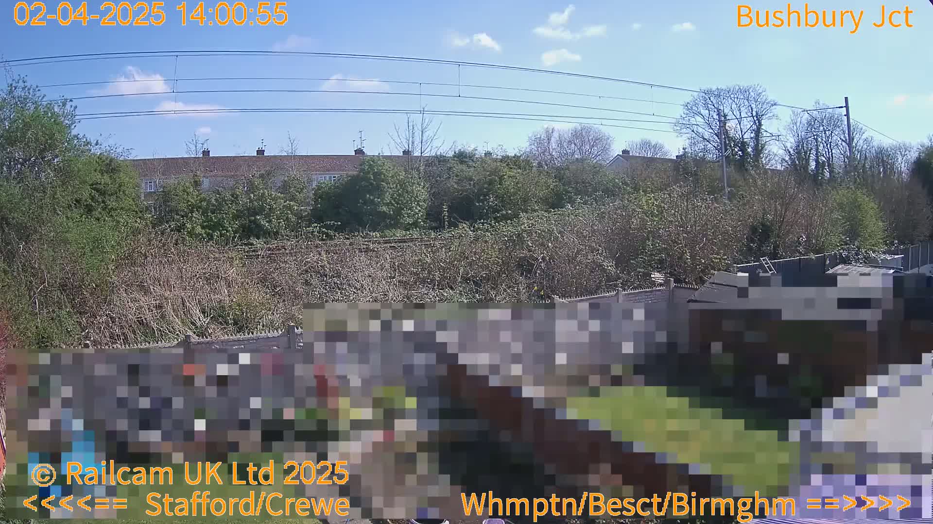 A sunny day shows a residential area with a backyard garden next to a railway line, partially obscured by overgrown vegetation.