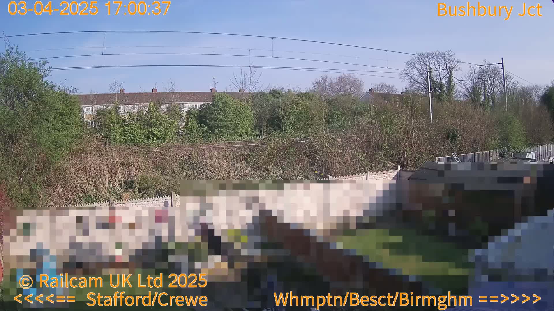 A sunny view of a backyard, featuring a wall, a garden, and railway lines and buildings in the distance.