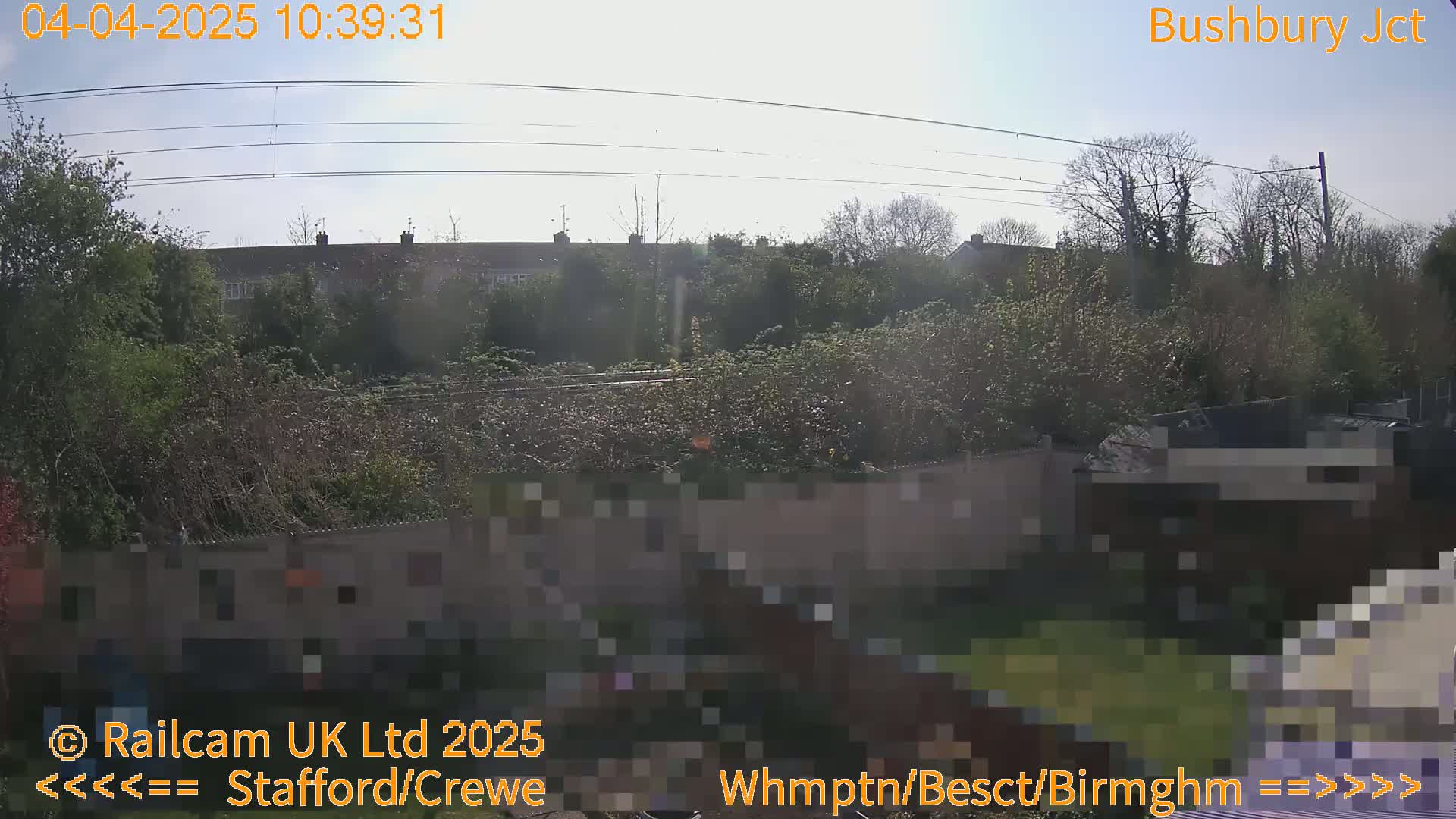 A sunny outdoor view shows a garden behind a fence, with railway lines and residential buildings visible in the background.