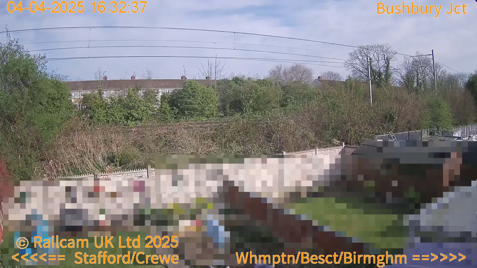 A backyard scene with a partially obscured shed and fence, overlooked by a row of houses and a railway line under a mostly sunny sky.