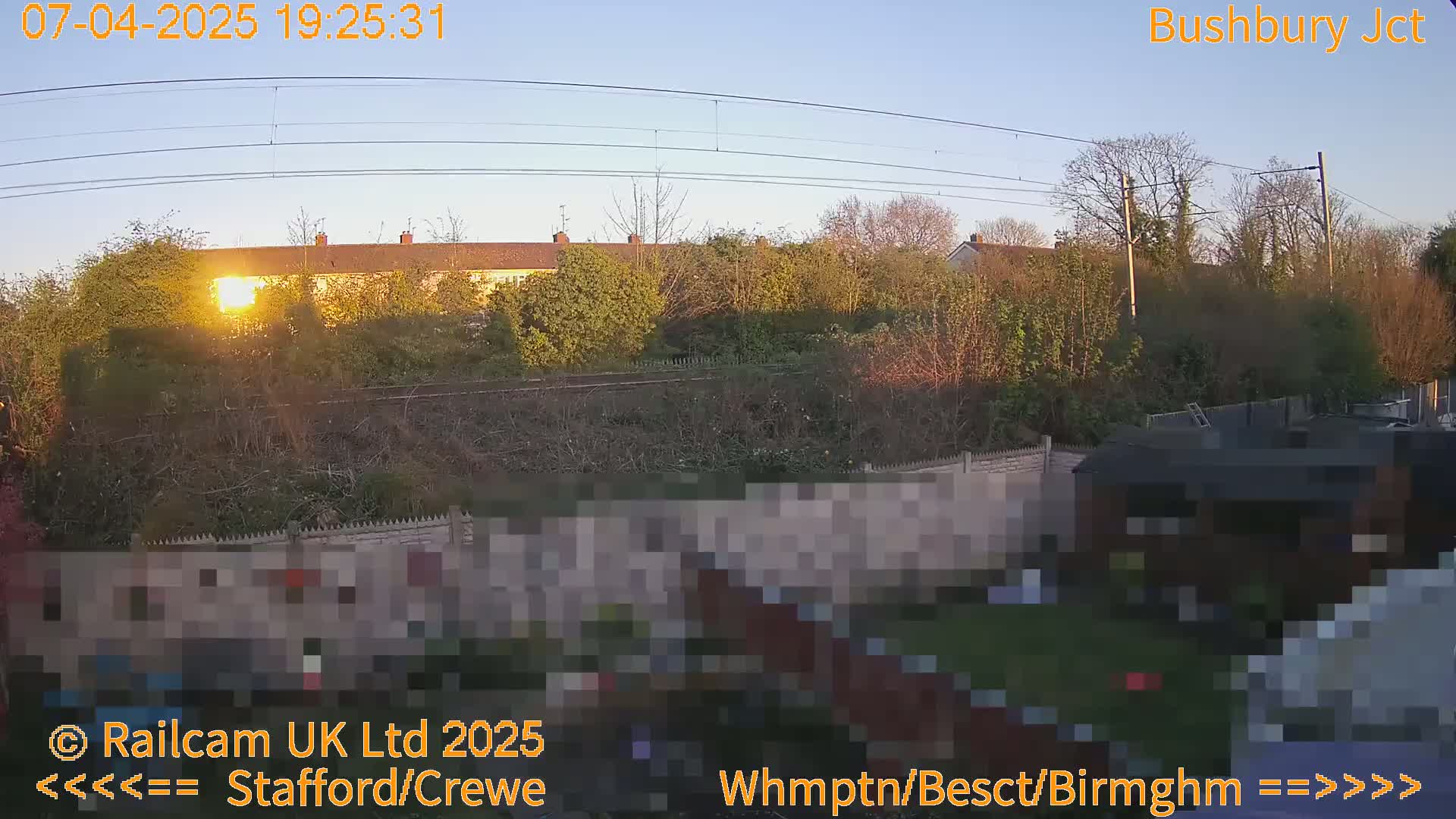 A low-hanging sun sets behind a line of houses and trees, casting long shadows over a backyard and railway tracks.
