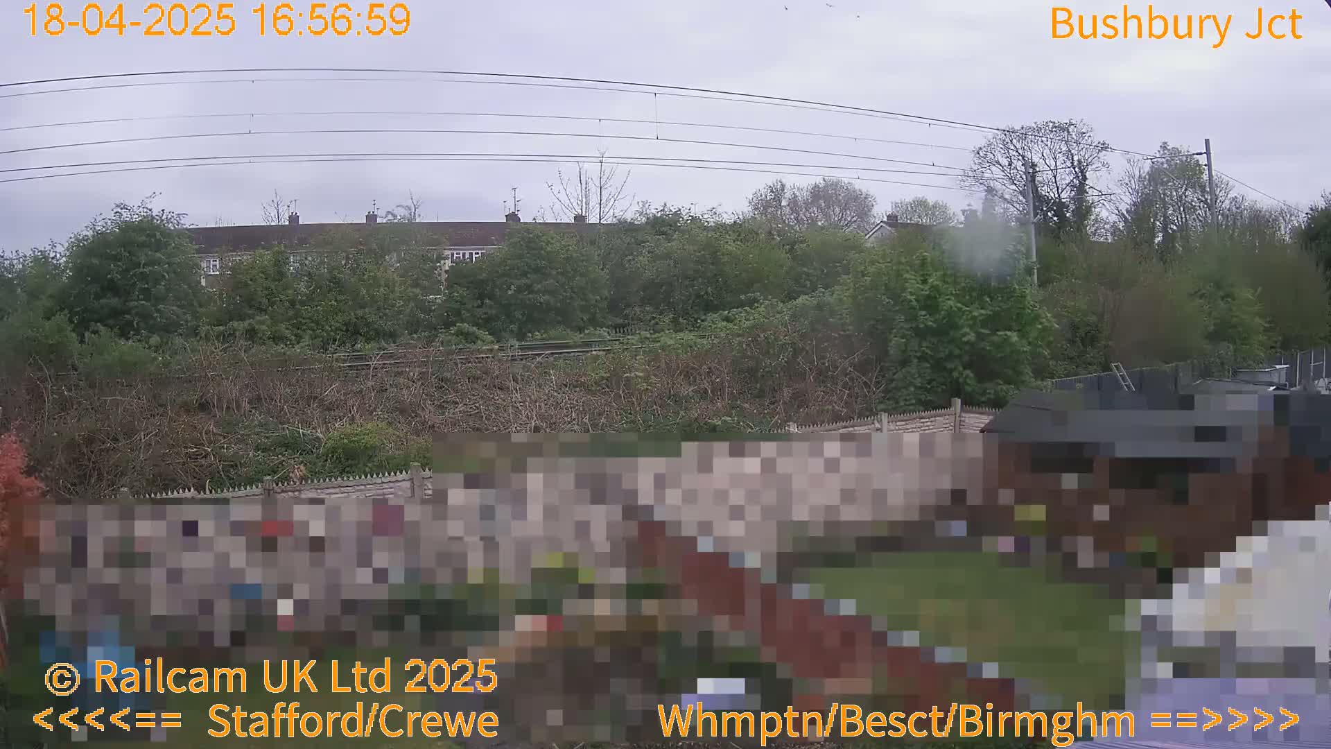 A view from a backyard shows a overcast sky, a garden, a fence, power lines, and houses and trees in the distance beyond a railway line.