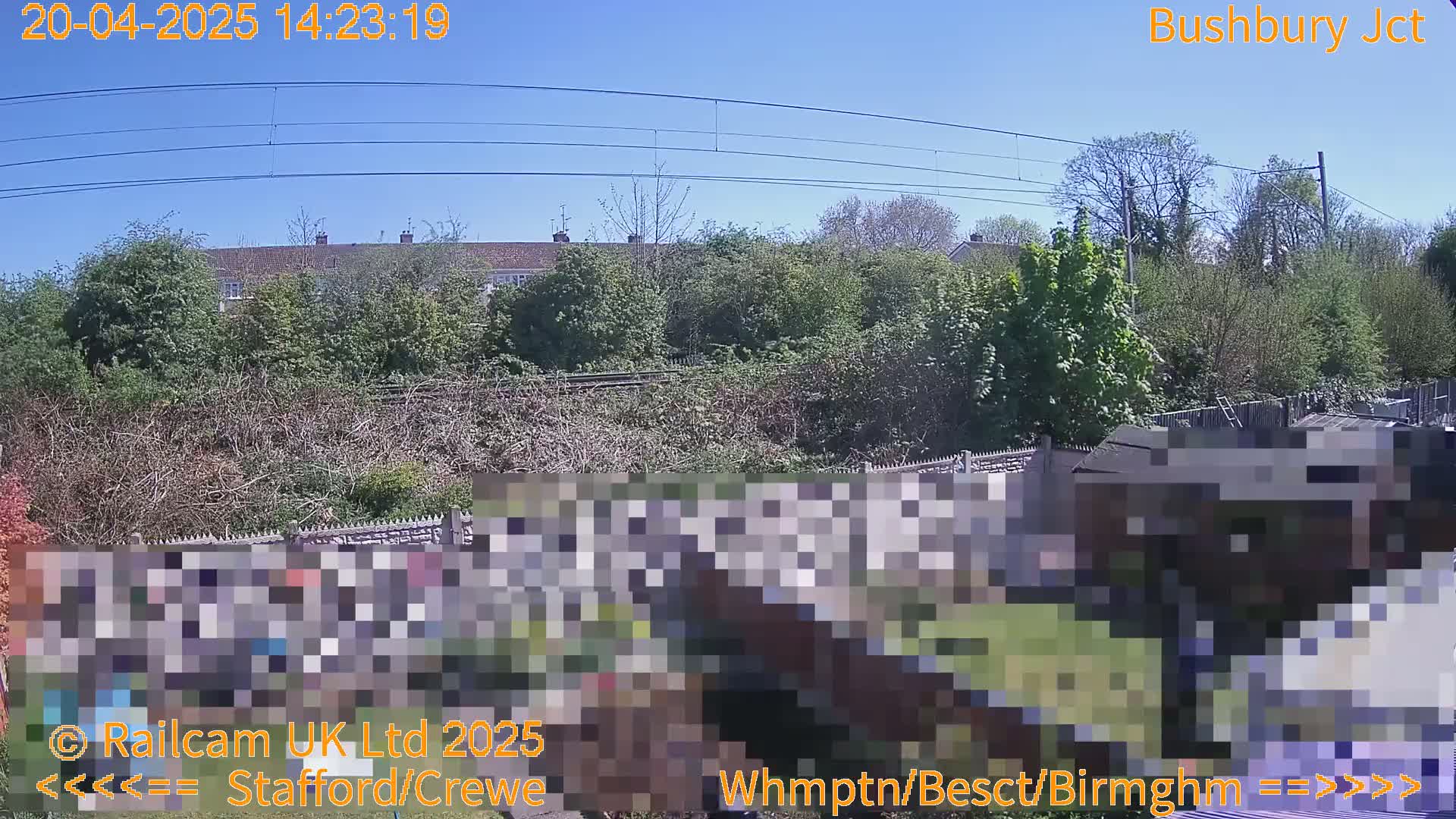 A sunny outdoor scene shows a railway line behind a row of houses and overgrown vegetation, with a partially obscured shed and garden in the foreground.