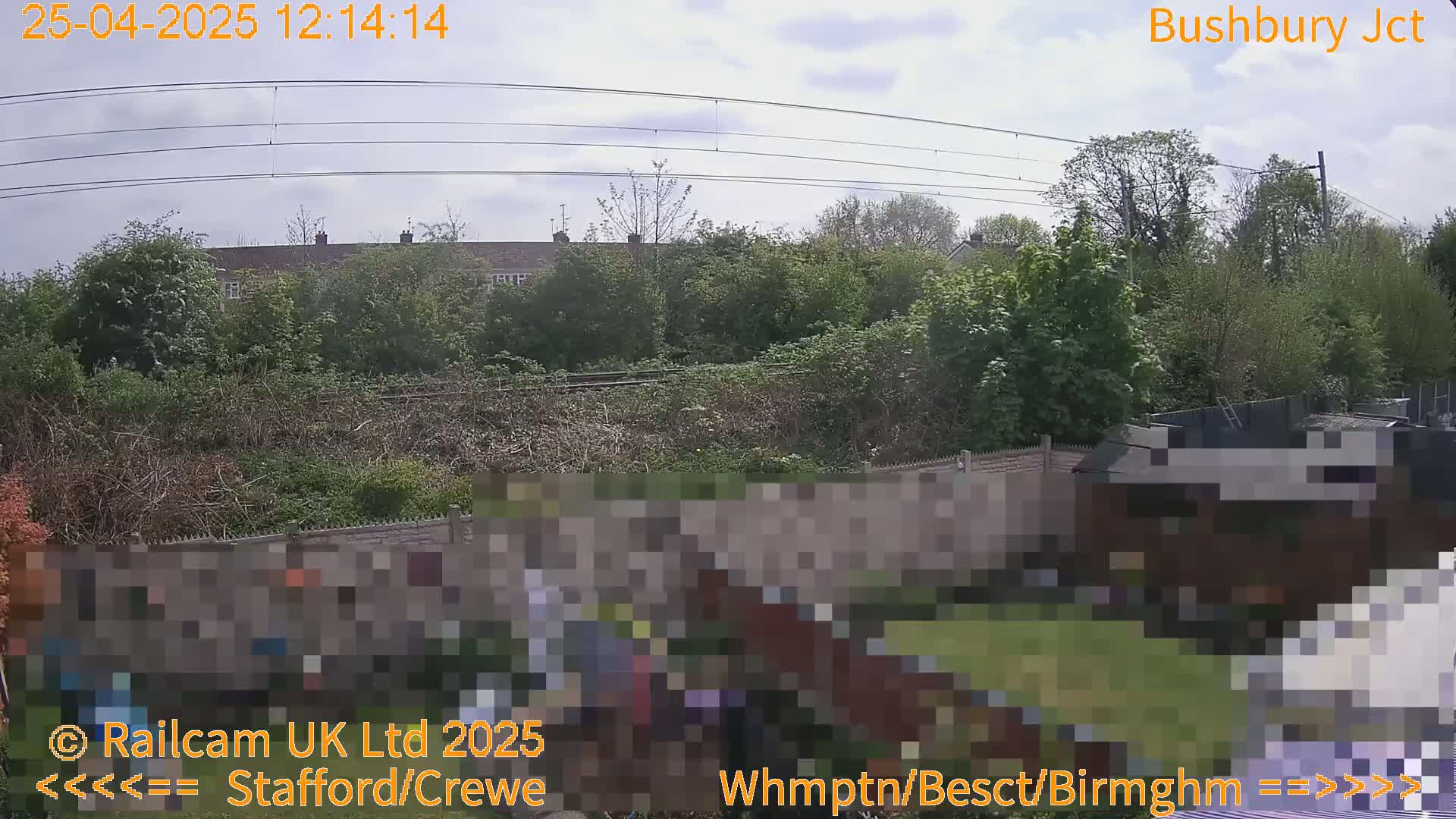 A partly cloudy day view of a backyard garden with a fence, trees, and houses in the background, beyond railway lines.