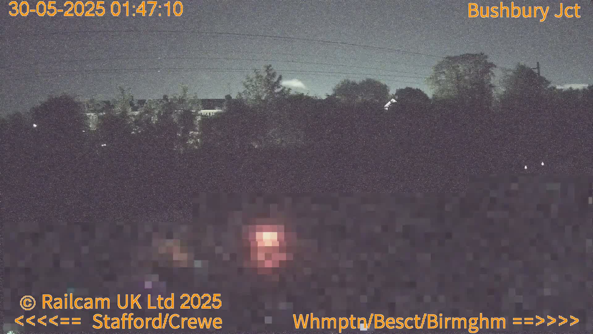 A nighttime, low-light view of trees and a low wall, possibly near a railway, under a dark, overcast sky.