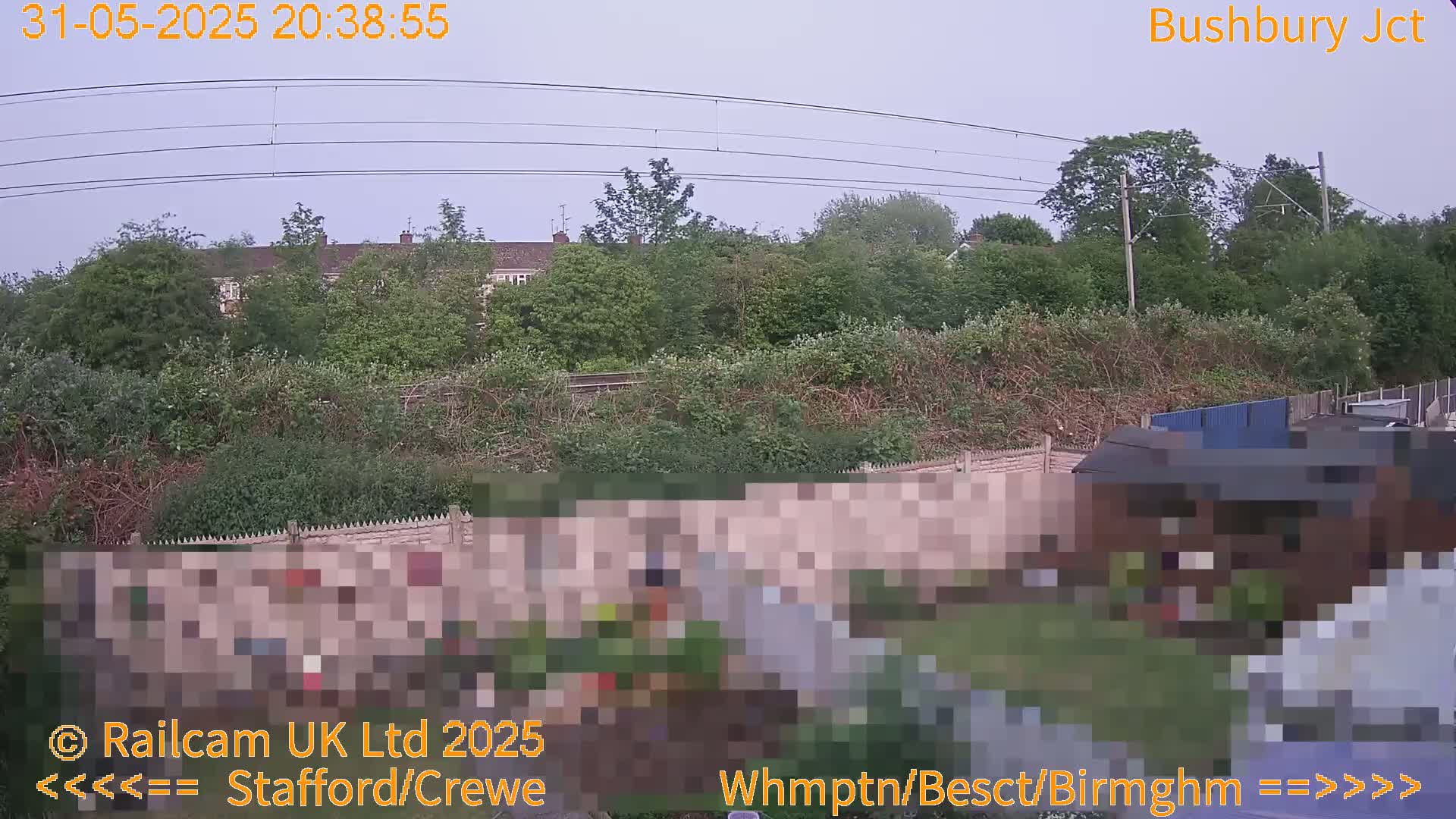 A view of a garden with a shed and a fence, overlooking a railway line and houses beyond, under an overcast sky.