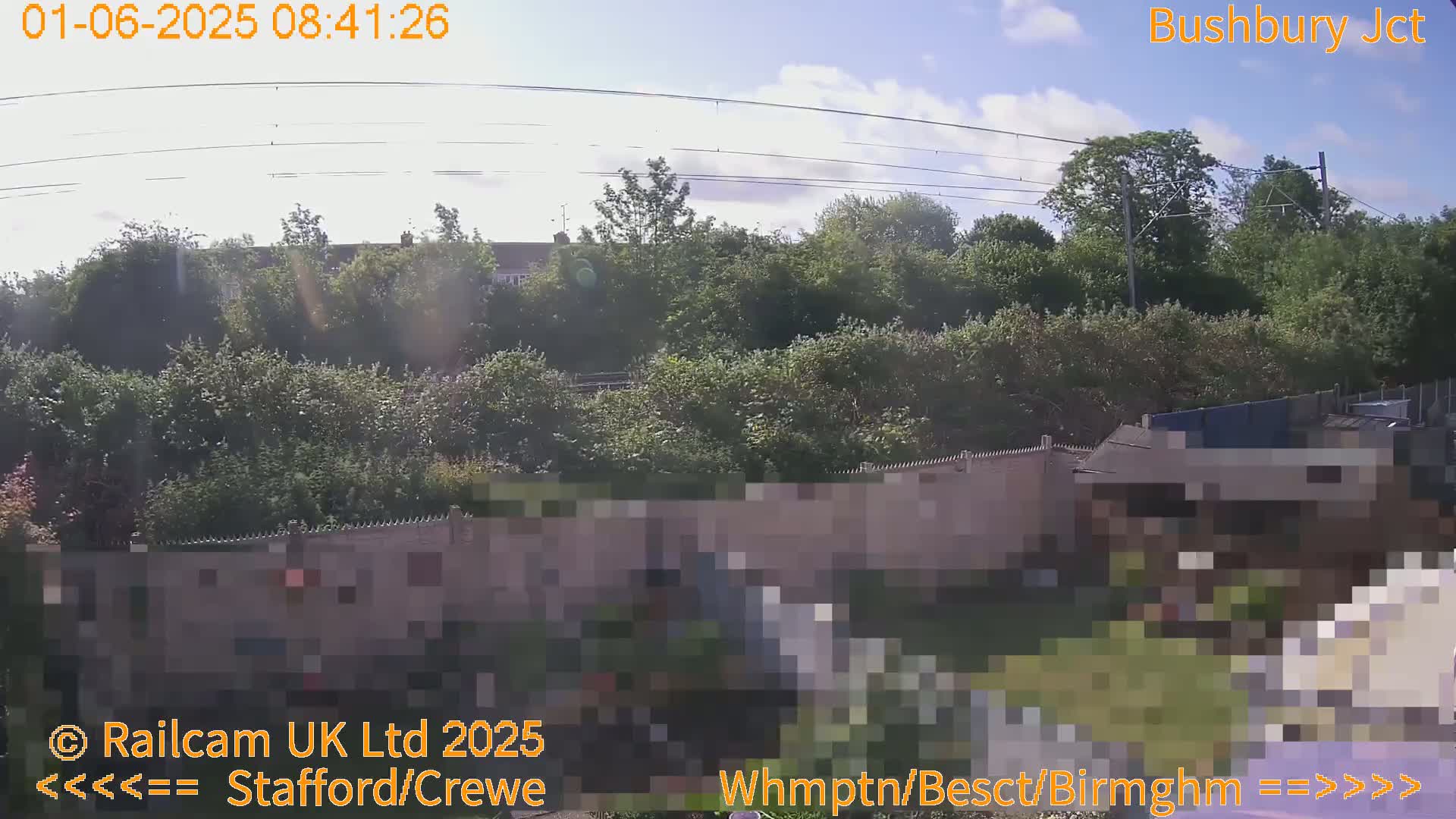 A mostly sunny day is shown overlooking a lush green landscape of trees and shrubs behind a wall, with power lines visible in the sky.
