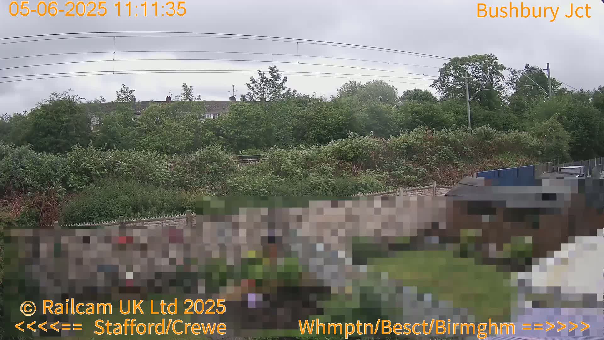 A view of a residential area on an overcast day, showing houses and lush green vegetation bordering a railway line with overhead power lines.