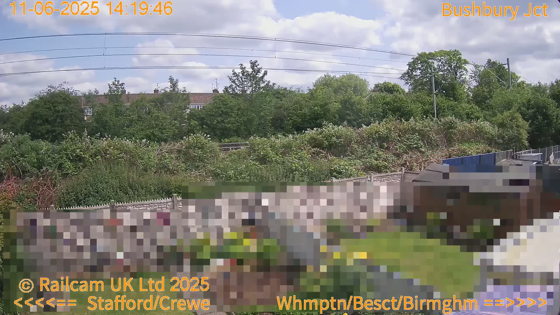 A partly sunny day shows a view of a garden next to a railway line, beyond which are houses and lush greenery.