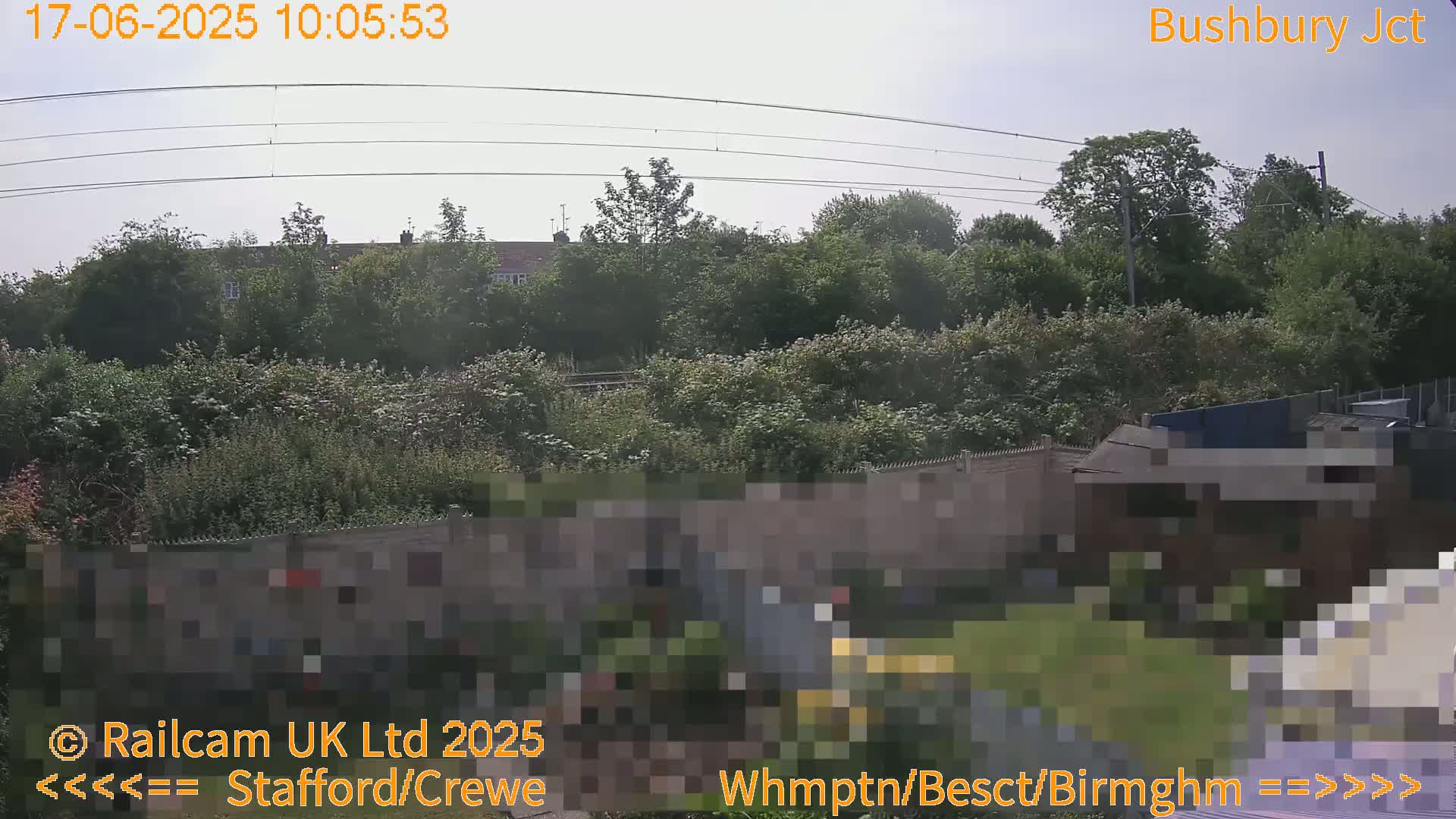 A mostly clear sky overlooks a lush green landscape with trees, houses, and a fence, partially obscuring a building.