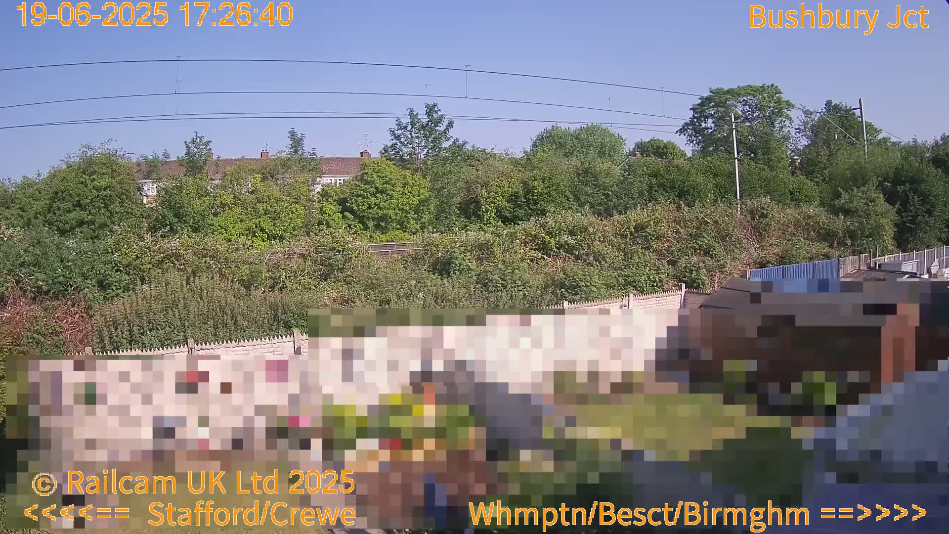 A clear, sunny day view of a residential area with lush green vegetation, fences, and a building partially obscured by pixelation.