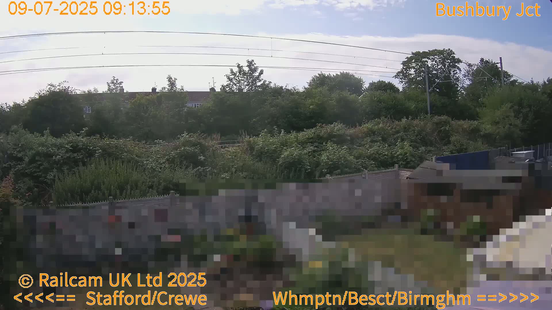 A partly cloudy sky overlooks a lush green backyard with a fence separating it from a railway line and houses beyond.