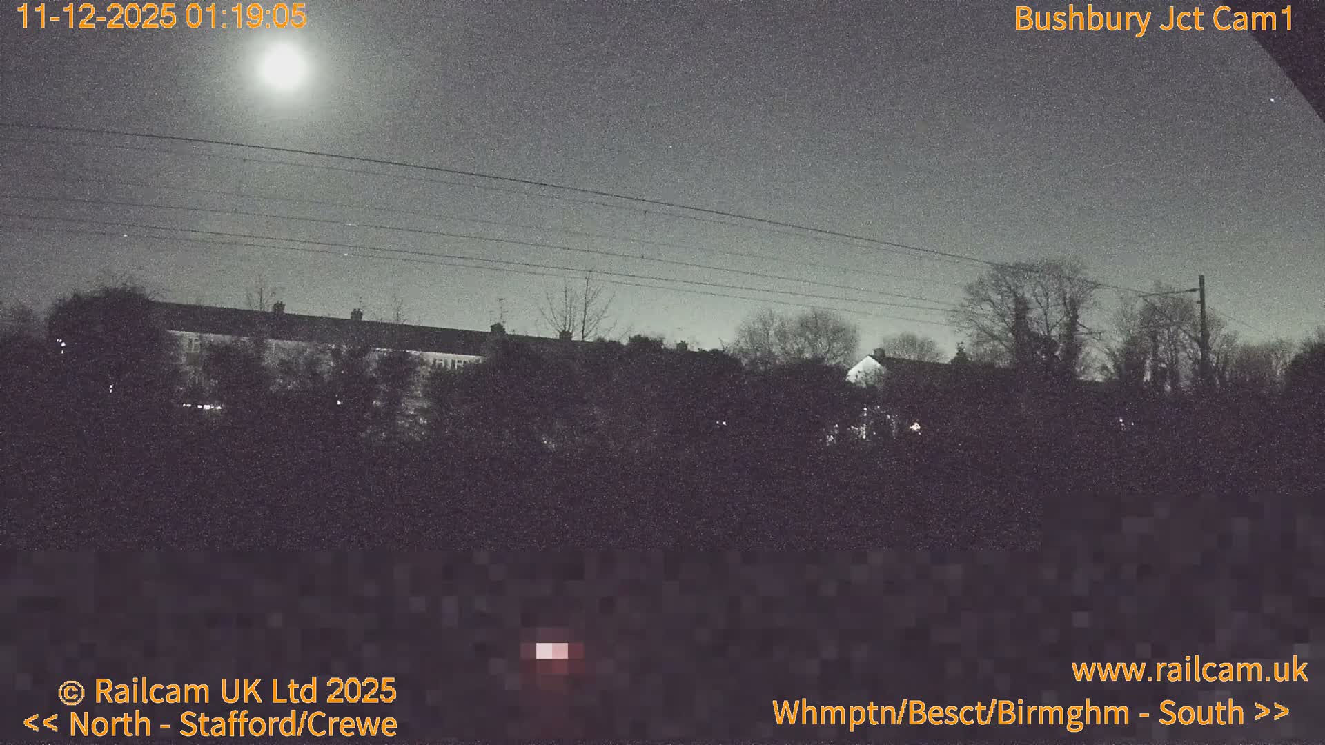 A grainy, low-light night view shows a bright moon in a clear sky above power lines, silhouetted trees, and distant houses.